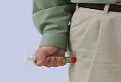 Una mano tieneе una siringa con liquido rosso vicino alla zona del gluteo di una persona che indossa una camicia verde e pantaloni chiari