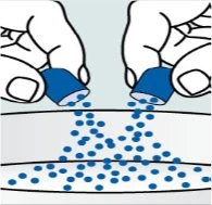 Two hands pouring small blue granules from two cylindrical containers onto a flat, light-colored surface