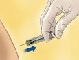 A gloved hand holding a syringe with a needle being inserted into the skin of the buttock at an angle of approximately 45 degrees, indicated by a blue arrow