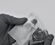 A gloved hand holding a transparent plastic package containing a medicine vial on a neutral grey background