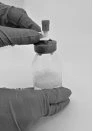 Two hands wearing protective gloves holding a glass vial containing a clear liquid with a dark stopper on a neutral background
