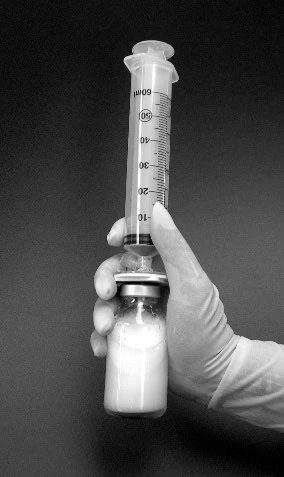 Ręka w rękawiczce trzyma pionowo strzykawkę 60 ml podłączoną do fiolki szklanej zawierającej mleczny biały płyn