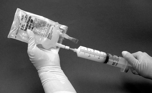 Ręce w rękawiczkach podłączają strzykawkę ze skalą pomiarową do worka z roztworem fizjologicznym chlorku sodu 0,9% w celu pobrania cieczy