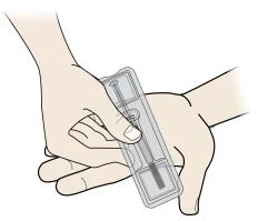 Dos manos sostienen y preparan un dispositivo médico rectangular transparente que contiene una jeringa con una aguja interna