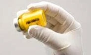 A hand wearing a white glove holding a vial with transparent yellow liquid against a light neutral background