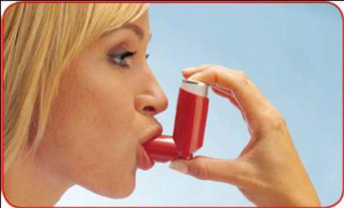 Una donna bionda tiene l'inalatore rosso alla bocca, inalando il farmaco contro un cielo azzurro chiaro