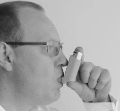 Un hombre con gafas sostiene el inhalador cerca de la boca, preparándose para inhalar la dosis del medicamento, en un primer plano en blanco y negro de perfil