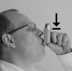 Un hombre con gafas sostiene el inhalador cerca de la boca, y una flecha negra indica la dirección de presión sobre el dispositivo durante la inhalación