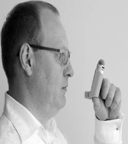 Un hombre con gafas y camisa blanca sostiene en la mano el inhalador en posición vertical frente al rostro para administrarse la dosis del medicamento