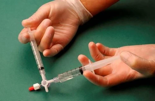 Дві руки в білих рукавицях тримають два шприци, з'єднані за допомогою білого з'єднувача з червоним клапаном на зеленому тлі