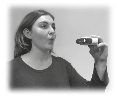 A woman in a dark sweater holding a small rectangular medical device in front of her face, looking at it with her mouth open