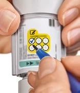 Manos sosteniendo un inyector blanco, con un bolígrafo azul marcando con una paloma azul uno de los seis recuadros en una tabla de control amarilla